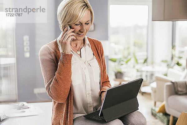 Unternehmerin spricht mit ihrem Smartphone  während sie zu Hause am Laptop arbeitet