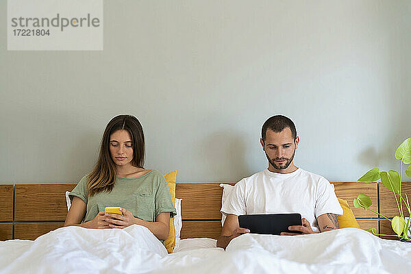 Junges Paar benutzt Smartphone und Tablet  während es zu Hause auf dem Bett sitzt