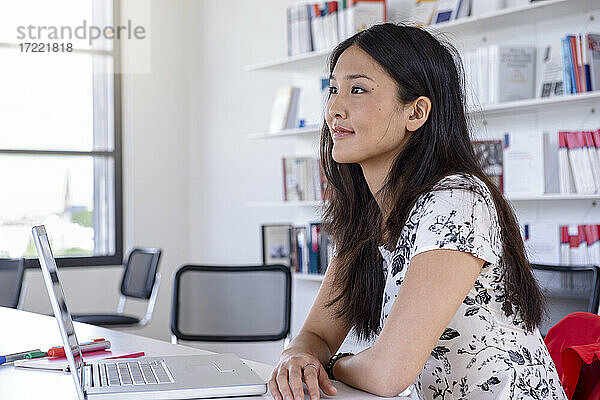 Nachdenkliche junge Frau sitzt mit Laptop am Schreibtisch in einer Bibliothek