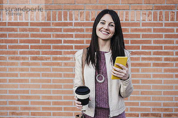 Lächelnde schöne Frau hält Smartphone und Einweg-Kaffeebecher vor einer Backsteinmauer