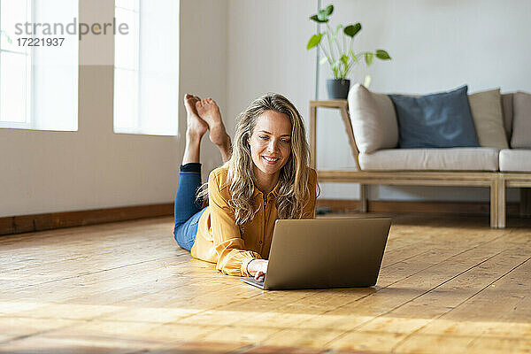 Lächelnde Frau  die einen Laptop benutzt  während sie auf dem Hartholzboden im Wohnzimmer liegt