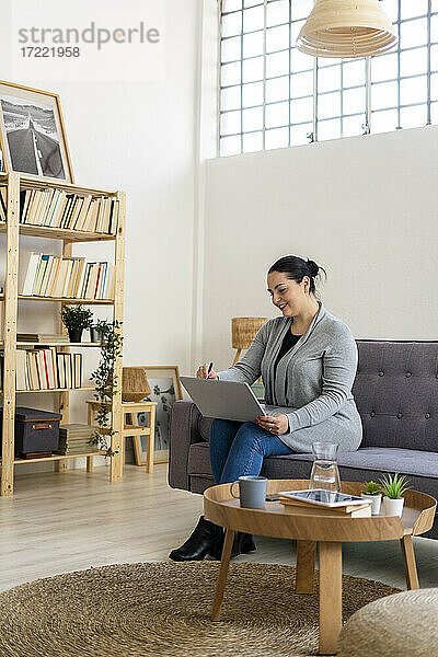 Lächelnde Frau  die einen Laptop benutzt  während sie zu Hause auf dem Sofa im Wohnzimmer sitzt