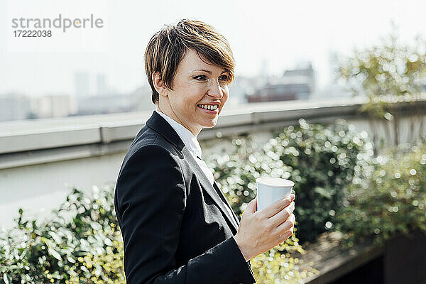 Lächelnde Unternehmerin  die eine Einweg-Kaffeetasse hält und wegschaut