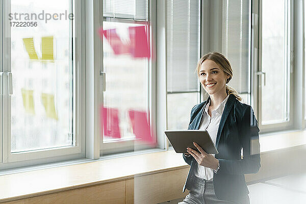 Zufriedene Geschäftsfrau mit digitalem Tablet und Blick auf Haftnotizen  die auf einem Glas im Büro kleben