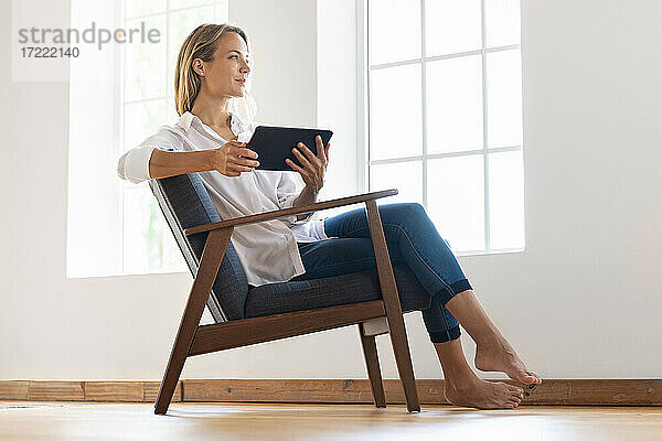 Nachdenkliche Frau  die ein digitales Tablet hält  während sie zu Hause auf einem Sessel sitzt