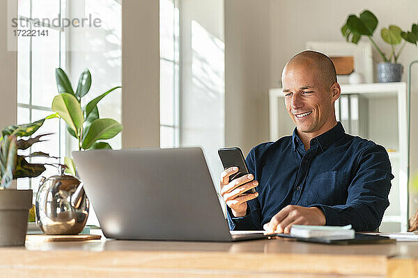 Lächelnder Geschäftsmann mit Laptop und Mobiltelefon bei der Arbeit zu Hause