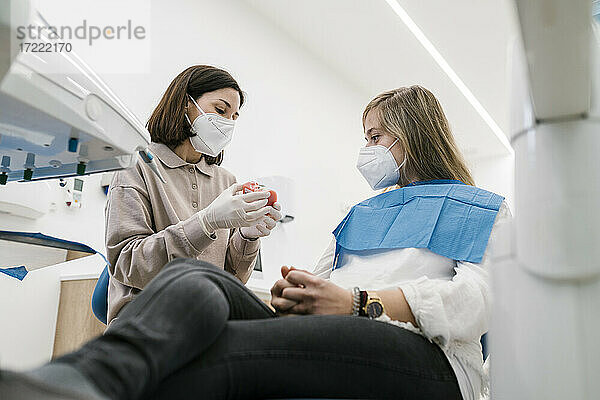 Weiblicher Kieferorthopäde mit Gesichtsmaske  der einem Patienten in der Praxis ein Modell künstlicher Zähne erklärt