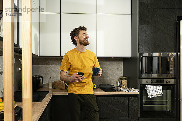 Glücklicher Mann mit Kaffeetasse und Smartphone in der heimischen Küche