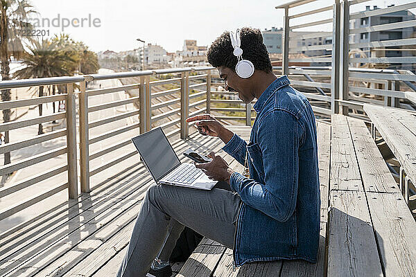 Fröhlicher Mann  der mit seinem Smartphone auf einen Laptop zeigt  während er auf einer Holztreppe an der Promenade sitzt