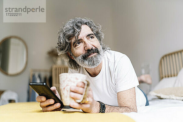 Mann mit Smartphone und Kaffeetasse schaut weg  während er zu Hause auf dem Bett liegt