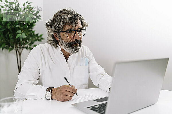 Seriöser Geschäftsmann schreibt auf Papier  während er im Büro am Laptop arbeitet