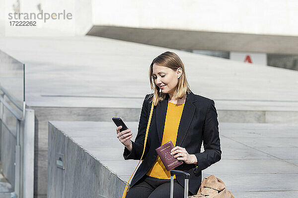 Unternehmer mit Reisepass  der ein Mobiltelefon benutzt  während er auf einer Stützmauer sitzt