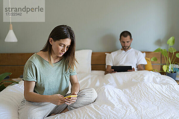 Junge Frau  die auf dem Bett ihr Smartphone benutzt  während ihr Freund im Hintergrund ein digitales Tablet betrachtet
