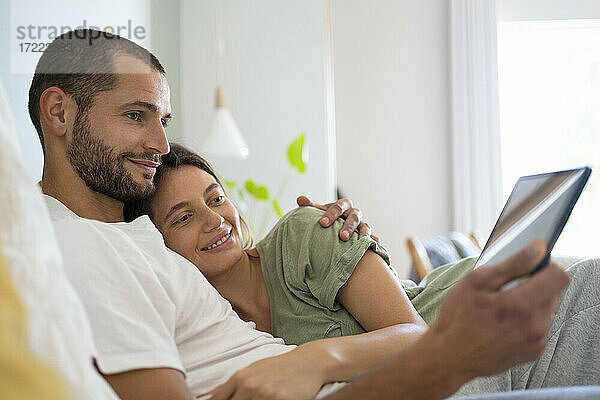 Junger Mann  der seine Freundin umarmt  während er zu Hause ein digitales Tablet benutzt