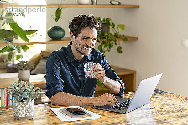 Lächelnder männlicher Freiberufler  der ein Glas Wasser in der Hand hält  während er im Heimbüro am Laptop arbeitet