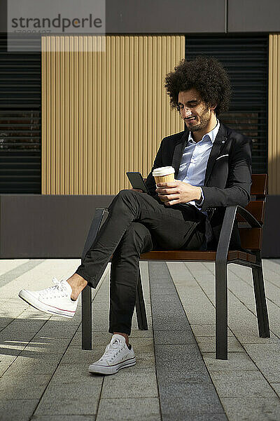Afro-Mann  der einen Kaffee trinkt und dabei sein Smartphone benutzt  während er auf einem Stuhl an einem sonnigen Tag sitzt