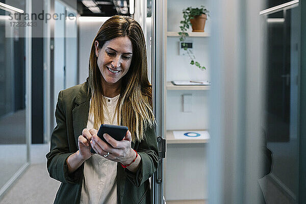 Lächelnde Frau  die ihr Smartphone benutzt  während sie sich an die Wand im Flur eines Büros lehnt