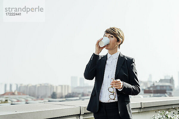 Weiblicher Geschäftsmann trinkt Kaffee  während er eine Brille an einer Stützmauer hält