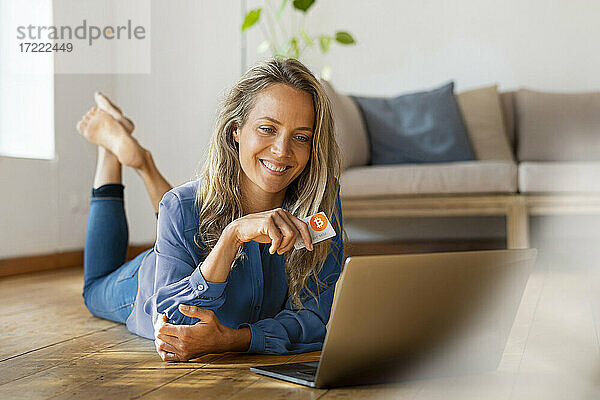 Lächelnde schöne Frau  die eine Kreditkarte in der Hand hält  während sie zu Hause vor einem Laptop auf dem Boden liegt