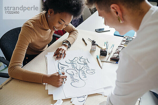 Lächelnde afrikanische Frau schreibt Kalligraphie auf Papier  während sie mit ihrem Freund im Atelier sitzt