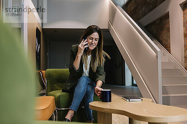 Geschäftsfrau  die mit einem Smartphone telefoniert und eine Kaffeetasse hält  während sie auf der Couch im Büro sitzt