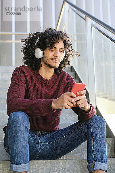 Junger gut aussehender Mann mit Kopfhörern  der sein Smartphone benutzt  während er auf einer Treppe sitzt