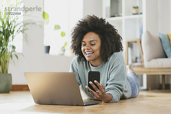 Lächelnde Frau mit Mobiltelefon und Laptop  während sie zu Hause auf dem Boden liegt