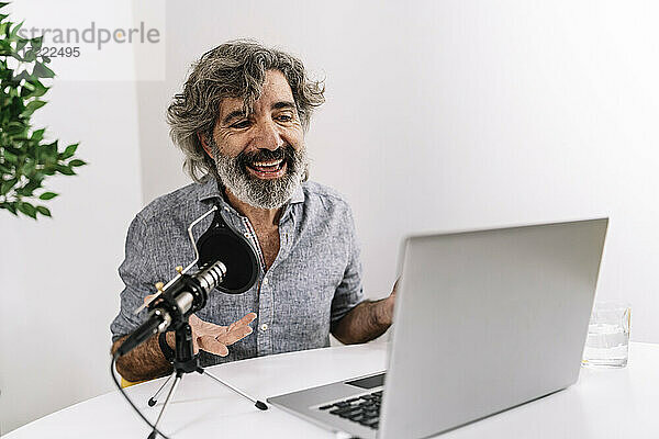 Lächelnder männlicher Podcaster  der vor einem Laptop auf einem Tisch im Büro sitzt
