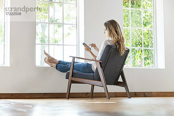 Entspannte Frau  die auf einem Stuhl zu Hause ihr Smartphone benutzt