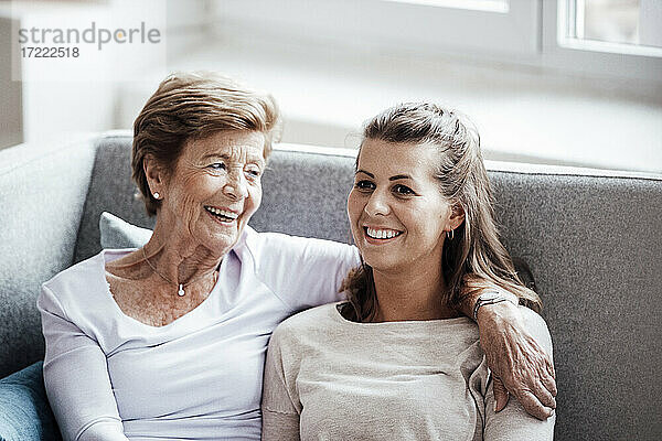 Lächelnde junge Frau  die den Arm um ihre Großmutter legt und wegschaut  während sie auf dem Sofa sitzt