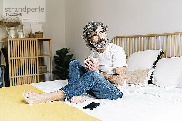 Nachdenklicher älterer Mann  der wegschaut und eine Kaffeetasse hält  während er zu Hause auf dem Bett sitzt