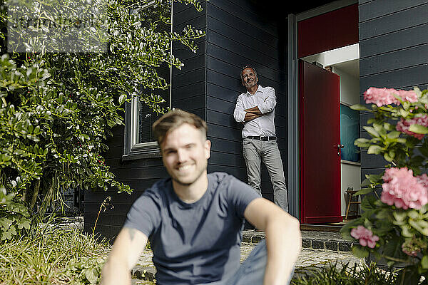 Lächelnder Vater lehnt sich an die Hauswand  während sein Sohn im Hinterhof sitzt