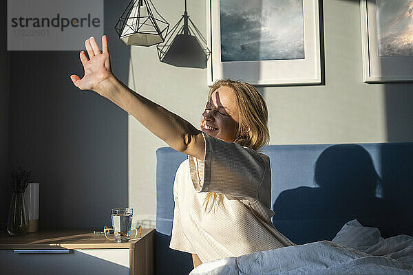 Lächelnde Frau streckt ihre Hand dem Sonnenlicht entgegen  während sie zu Hause auf dem Bett sitzt