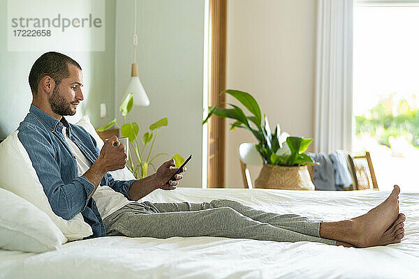 Junger Mann mit Kaffeetasse und Smartphone zu Hause