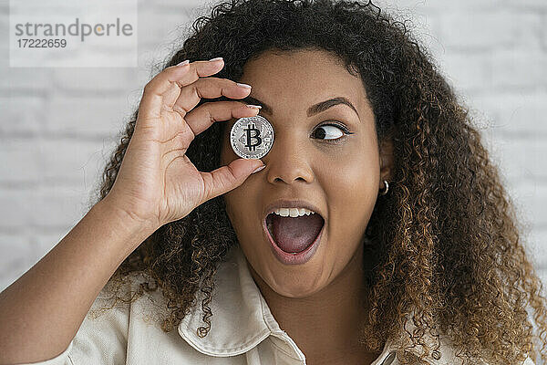 Aufgeregte Frau mit offenem Mund  die ihre Augen mit Bitcoin vor einer Wand bedeckt