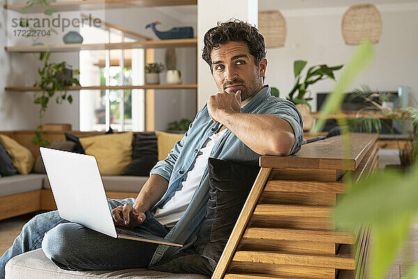 Nachdenklicher männlicher Freiberufler  der mit seinem Laptop auf dem Sofa sitzt und wegschaut