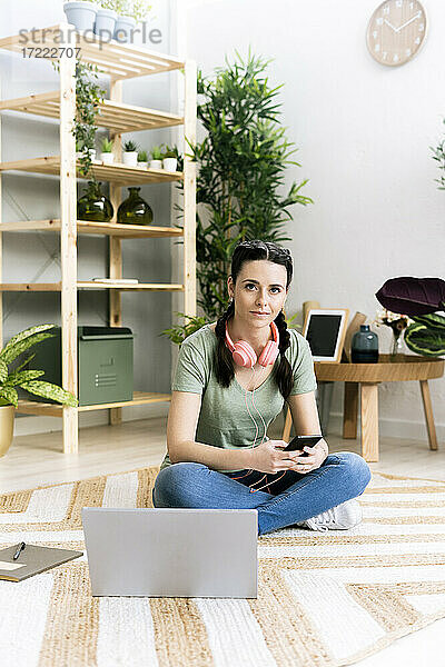 Frau mit Smartphone sitzt zu Hause am Laptop