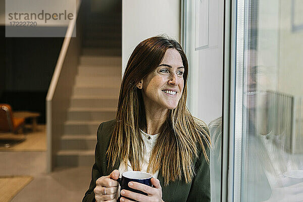 Nachdenkliche Geschäftsfrau mit Kaffeetasse  die durch ein Fenster im Büro schaut