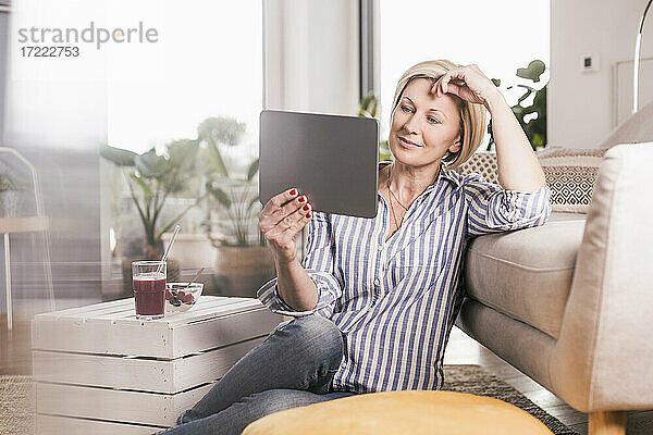 Ältere Frau schaut auf ein digitales Tablet  während sie sich zu Hause am Sofa anlehnt
