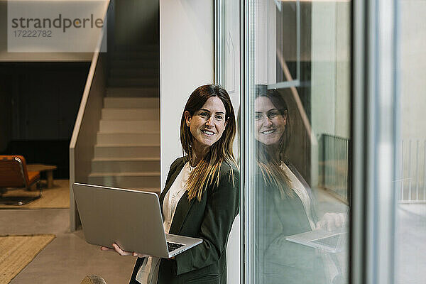 Nachdenkliche Geschäftsfrau  die einen Laptop hält und durch ein Glasfenster im Büro schaut
