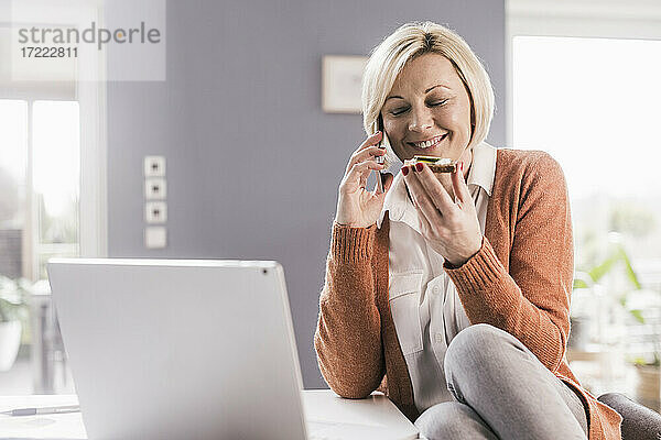 Lächelnde Geschäftsfrau mit geschlossenen Augen  die Brot isst  während sie mit dem Handy im Büro zu Hause telefoniert