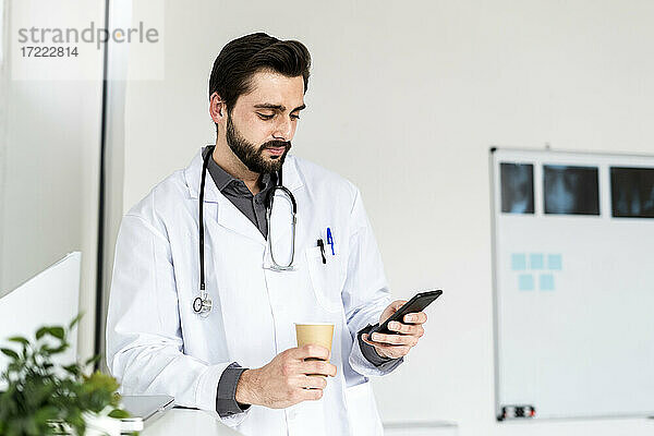 Männlicher Arzt  der eine Einweg-Kaffeetasse hält  während er sein Smartphone im Krankenhaus benutzt