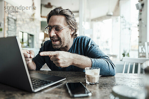 Lächelnder Mann gestikuliert während eines Videoanrufs über einen Laptop in einem Café