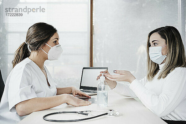 Patient nimmt Desinfektionsmittel ein  während er mit einer Ärztin am Schreibtisch im Krankenhaus während der Pandemie spricht