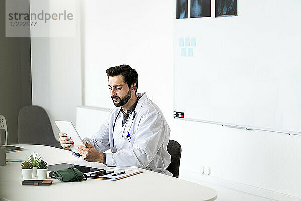 Gutaussehender männlicher Arzt  der ein Tablet benutzt und am Schreibtisch im Krankenhaus sitzt