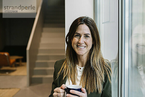 Lächelnde Geschäftsfrau mit Kaffeetasse  die sich an ein Glasfenster im Büro lehnt