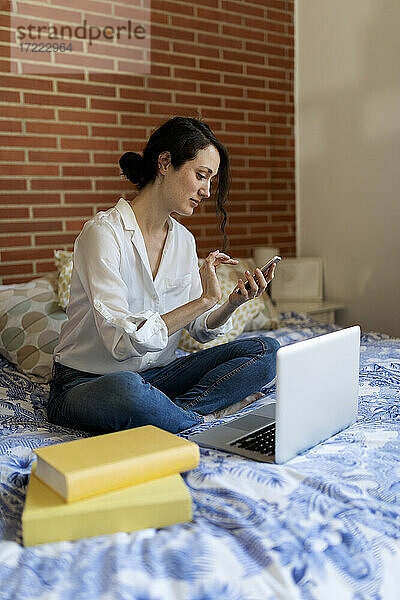 Frau  die ein Mobiltelefon benutzt  während sie zu Hause im Schlafzimmer am Laptop sitzt
