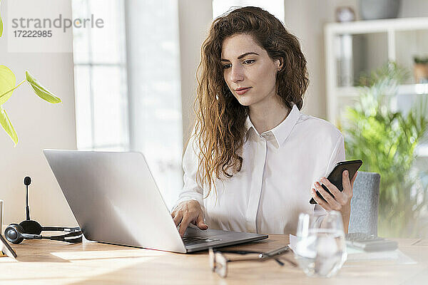 Berufstätige Frau mit Mobiltelefon  die an einem Laptop am Schreibtisch arbeitet