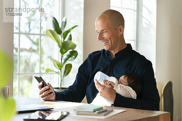 Vater füttert ein kleines Mädchen mit Milch  während er im Büro zu Hause am Smartphone arbeitet