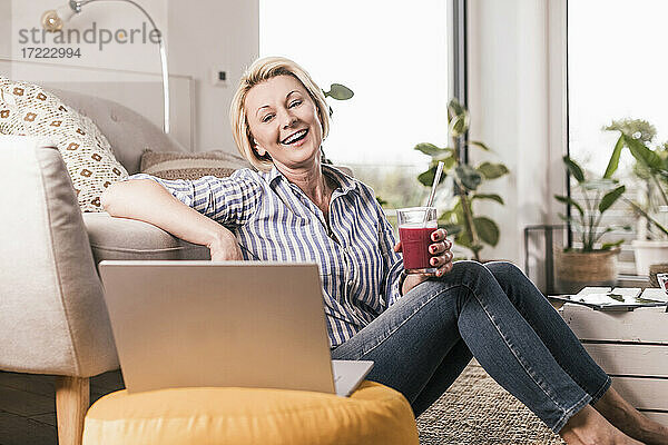 Glückliche reife Frau  die ein Saftglas hält  während sie zu Hause auf den Laptop schaut